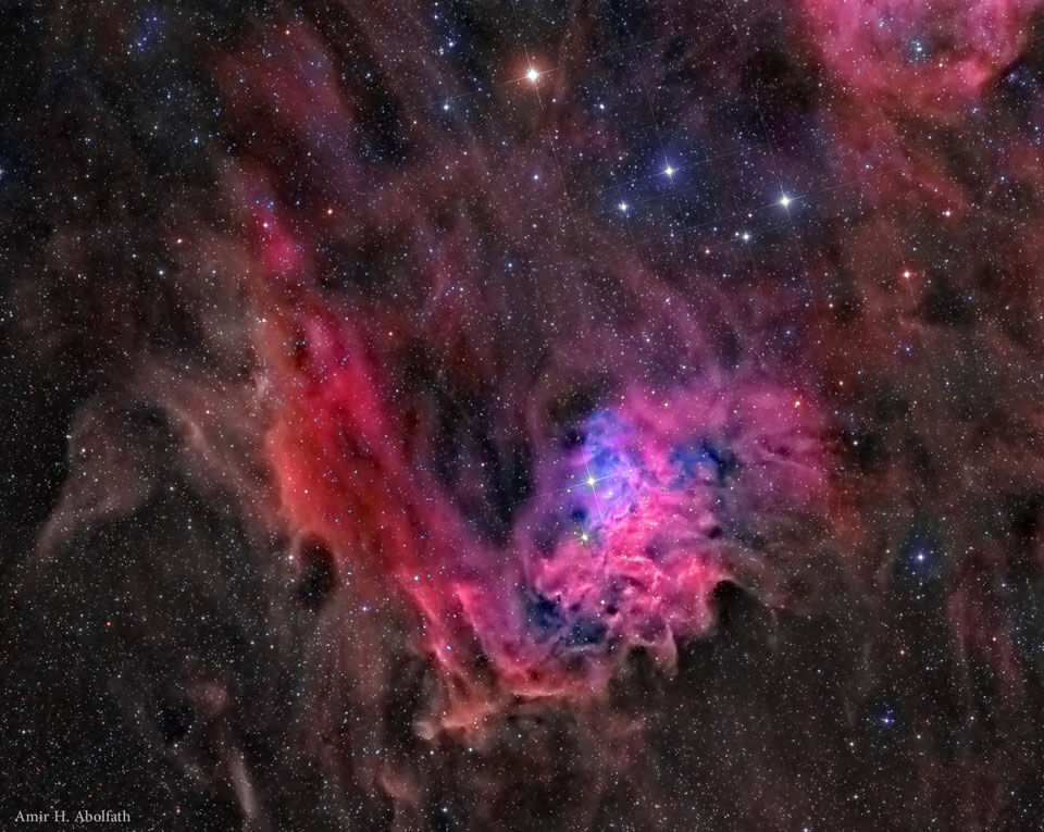 Das Bild ist von rötlichen und braunen Nebelfetzen gefüllt, die von Sternen gesprenkelt sind. In der Mitte ist eine stark strukturierte, heller leuchtende zyklamefarbene Ballung.