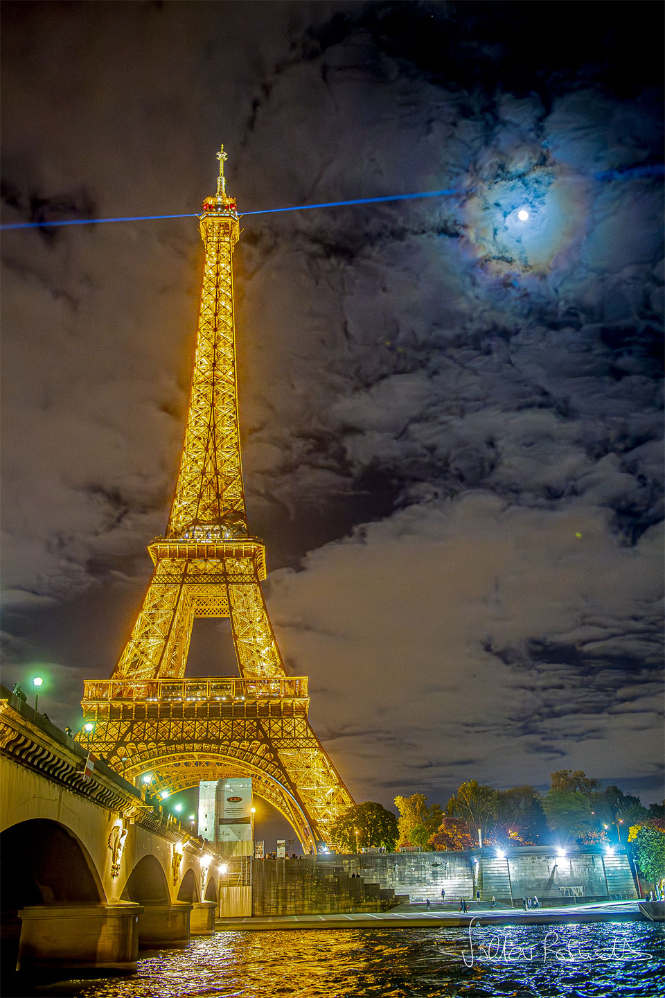 Links ragt der gelb beleuchtete Eiffelturm hinter einer Brücke auf, oben schießt scheinbar ein blauer Strahl zum Mond, der von einem blau leuchtenden Hof mit rötlichem Ring umgeben ist.