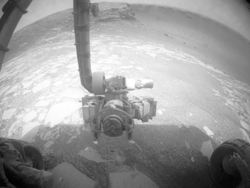 Opportunity blickt auf den Mars. Von oben ragt ein Arm mit Instrumenten ins Bild. Links und rechts sind unten die Räder des Rovers zu sehen. Das Schwarzweiß-Bild entstand mit einer Weitwinkelkamera, die Hindernisse erkennen soll.