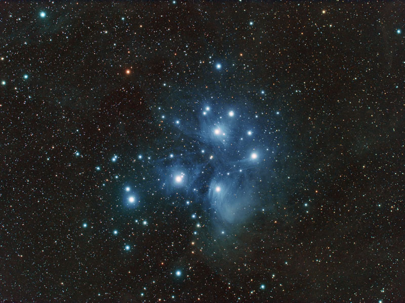 Mitten im Bild ist ein Sternhaufen, dessen Sterne von blauen Reflexionsnebeln umgeben sind.