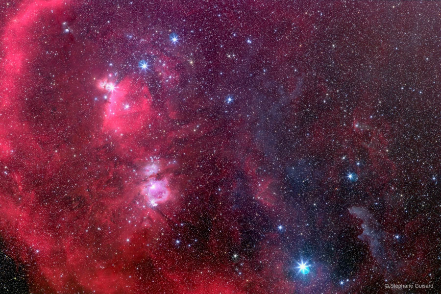 Um den Gürtel des Orion ist eine riesige Molekülwolke mit vielen Strukturen, die im Bild als rötlich leuchtende Nebel zu sehen sind, nur der Hexenkopfnebel rechts unten leuchtet bläulich.
