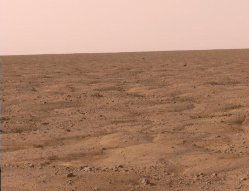 Der Boden im Vordergrund ist rötlich und wirkt sandbedeckt, der Himmel hinter dem Horizont ist rosarot.