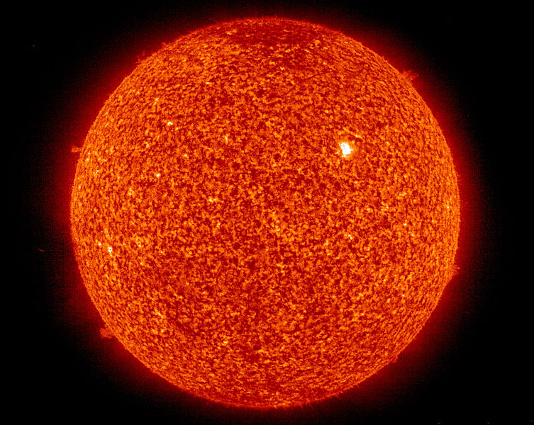 Das Bild zeigt die Sonne vor schwarzem Hintergrund in einer rötlichen Farbe, welche die Granulation zeigt. Am Rand sind einige Protuberanzen zu sehen.
