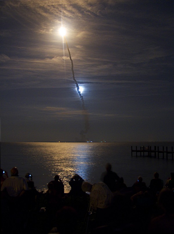Hinter einem Gewässer startet eine Rakete. Das Feuer der Triebwerke spiegelt sich im Wasser, hinter der Abgasfahne leuchtet der Mond.