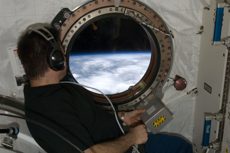 Ein Astronaut mit Kopfhörern blickt aus einem runden Fenster, durch das man den Rand der Erde sieht.