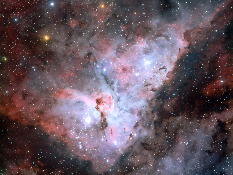 Das Bild enthält dünn verteilte Sterne und ist mit Nebeln gefüllt. In der Mitte leuchten rötliche und hellviolette Nebel, an den Rändern befinden sich Dunkelnebel.