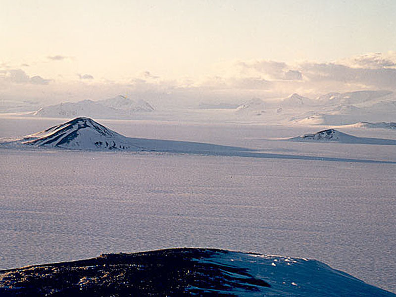 Die sonnige Landschaft ist mit Eis bedeckt, aus dem einige Gipfel ragen.