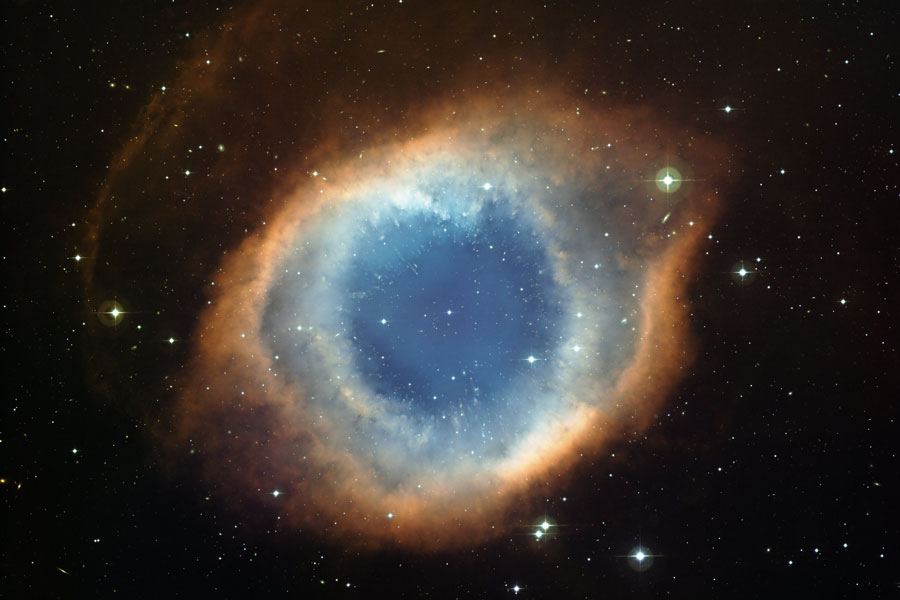 In der Mitte leuchtet ein ringförmiger Nebel mit einem bräunlich-orangefarbenen Rand, der nach innen hin weißlich wird und in der Mitte dunkelblau leuchtet.