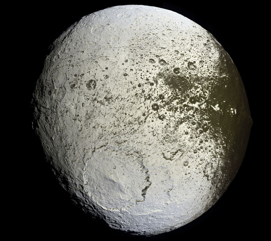 Der weiße Mond Iapetus hat unten einen sehr großen Krater. Die rechte Seite ist von einer dunklen Substanz überzogen.