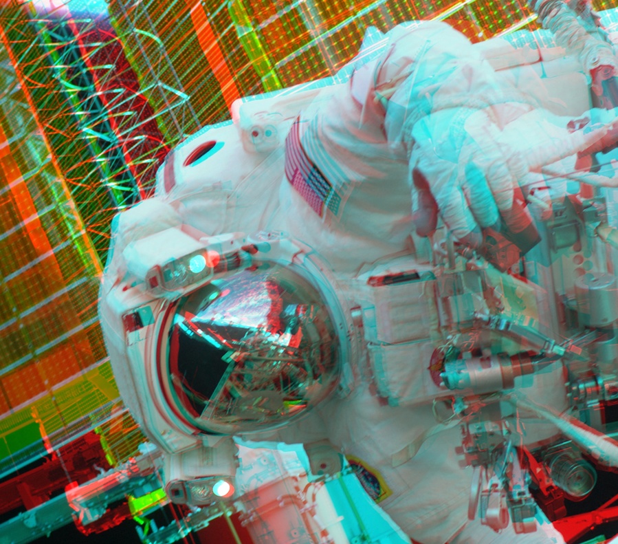 Von rechts ragt schräg ein Astronaut (Kopf und Rumpf) ins Bild, er ist vor einem gelb-roten Hintergrund aus Paneelen abgebildet. Mit rot-blauen Brillen wirkt das Bild dreidimensional.