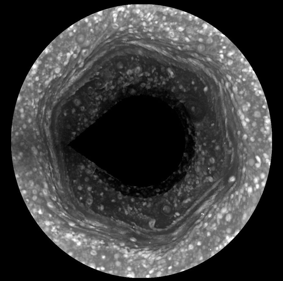 Der von einem schwarzen Rahmen umgebene Kreis zeigt eine sechseckige Struktur bei Saturns Nordpol. In der Mitte ist ein dunkler Kreis, darum herum ist eine sechseckige Struktur, alle hellen Bereiche sind von vielen hellen runden Flecken gesprenkelt. 