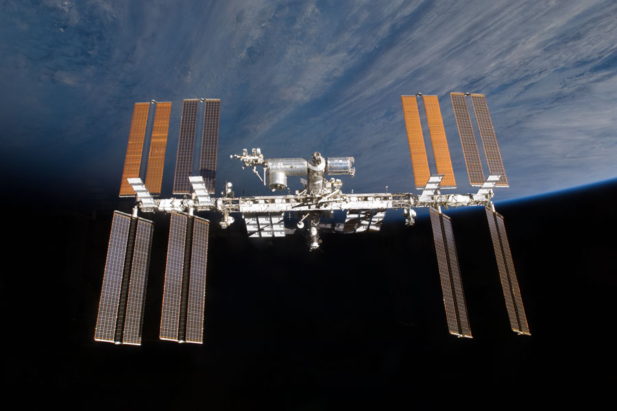 Im Bild schwebt die Internationale Raumstation. Links und rechts ragen je 4 Solarpaneele nach oben und nach unten. Im Hintergrund ragt nach oben die Erde auf.