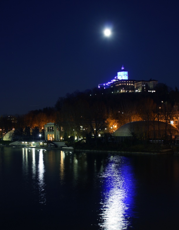 Hinter dem italienischen Fluss Po ragt ein Berg auf, oben steht die Kirche von Turin, darüber leuchtet der Mond, der sich weiß und blau im Wasser spiegelt. 
