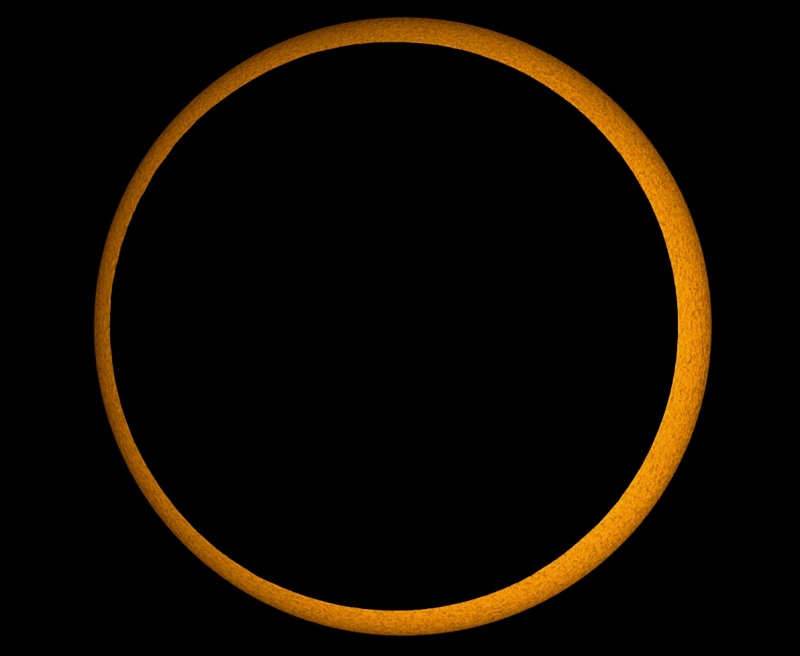 Ein schwarzer Kreis mitten im Bild ist von einem hellen orangefarbenen Ring umgeben, der wiederum einen schwarzen Hintergrund hat. Der schwarze Kreis ist der Mond, der orangefarbene Ring die Sonne.