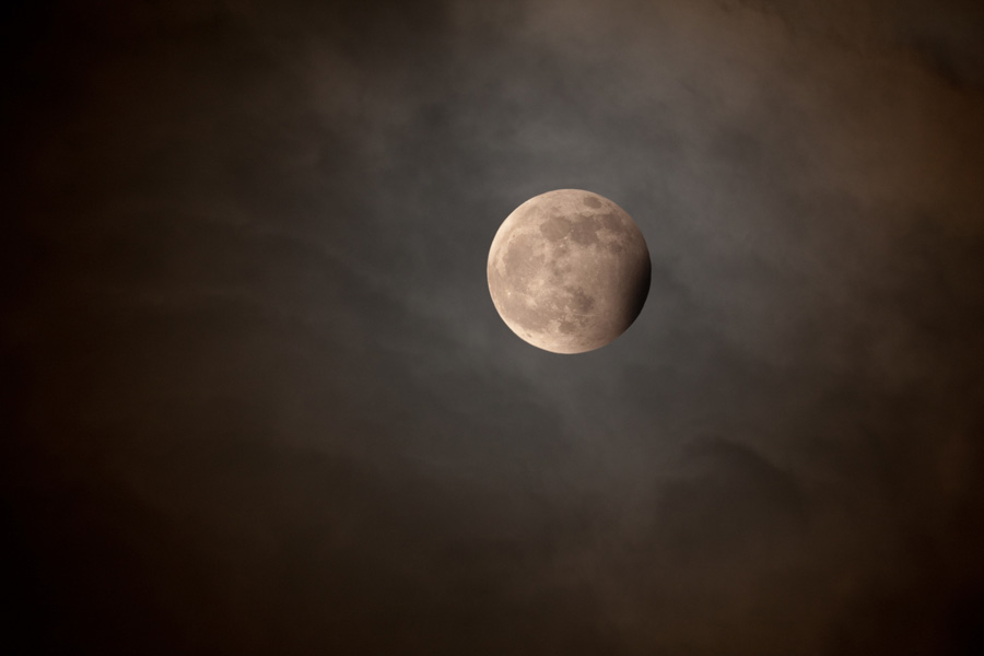 An einem wolkig wirkenden Himmel schwebt ein sehr plastischer Mond mit dunklen Meeren, der rechts unten einen dunklen Schatten hat.