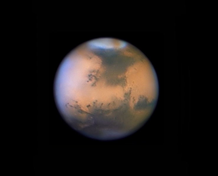 Im Bild ist der Planet Mars vor dunklem Hintergrund zu sehen, de Rand wirkt eher verschwommen, oben ist eine helle Polkappe, über der eine rote Wolke verläuft, die dunklen Strukturen auf der Oberfläche wirken zum Teil sehr klar.