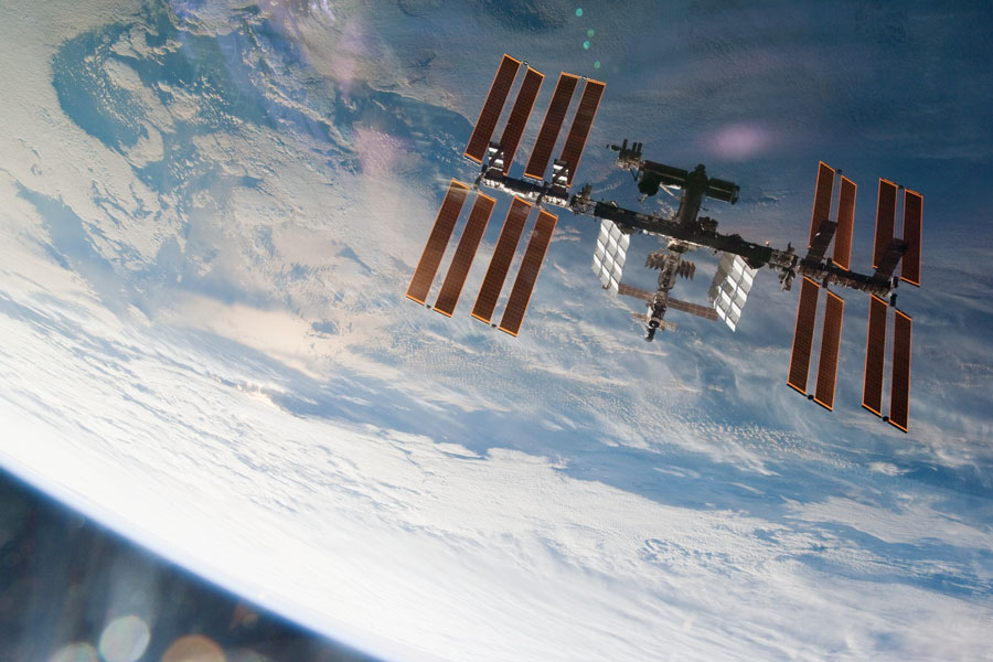 Vor der Erde, die von oben ins Bild ragt, schwebt die Internationale Raumstation mit mächtigen Solarpaneelen. Links unten ist in der Ecke der Erdrand zu sehen.