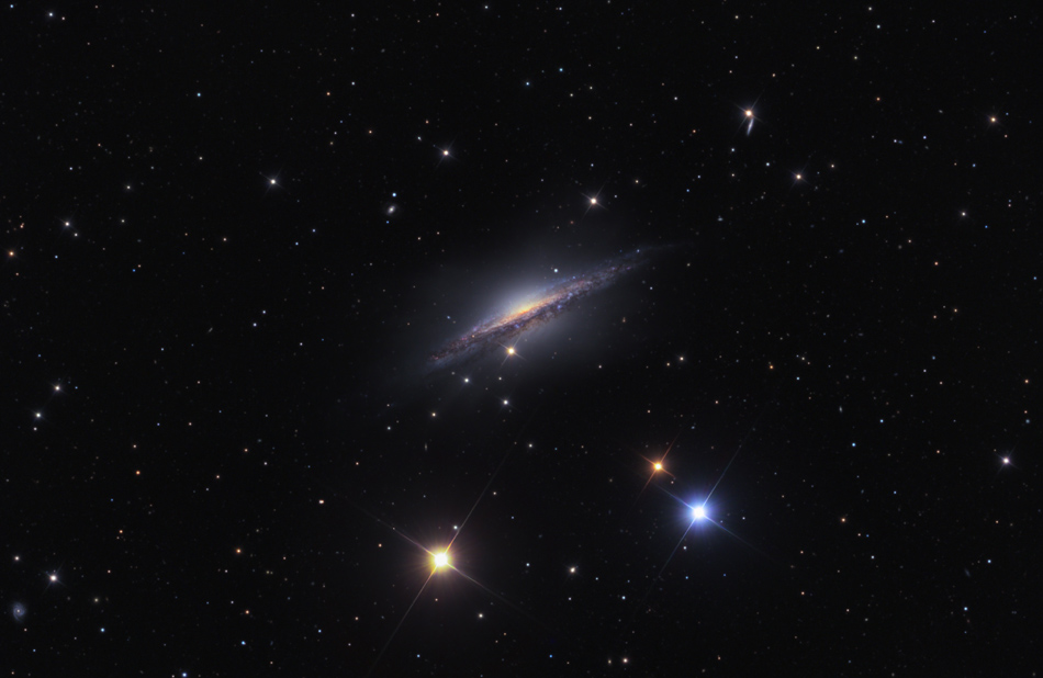 Mitten im Bild leuchtet eine kleine Spiralgalaxie. Wir blicken auf die Kante, die von einem breiten Staubband durchzogen ist. Darunter sind zwei markante gezackte Sterne, links ein gelber, rechts ein blauer.