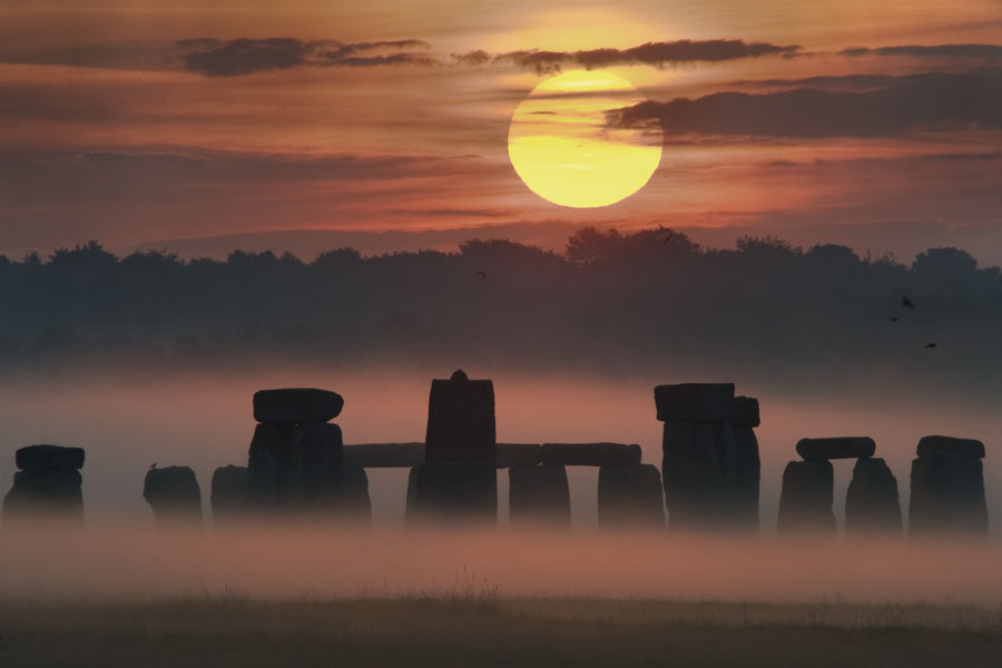 Über Stonehenge im Morgennebel geht die Sonne an einem leicht wolkigen Himmel auf.