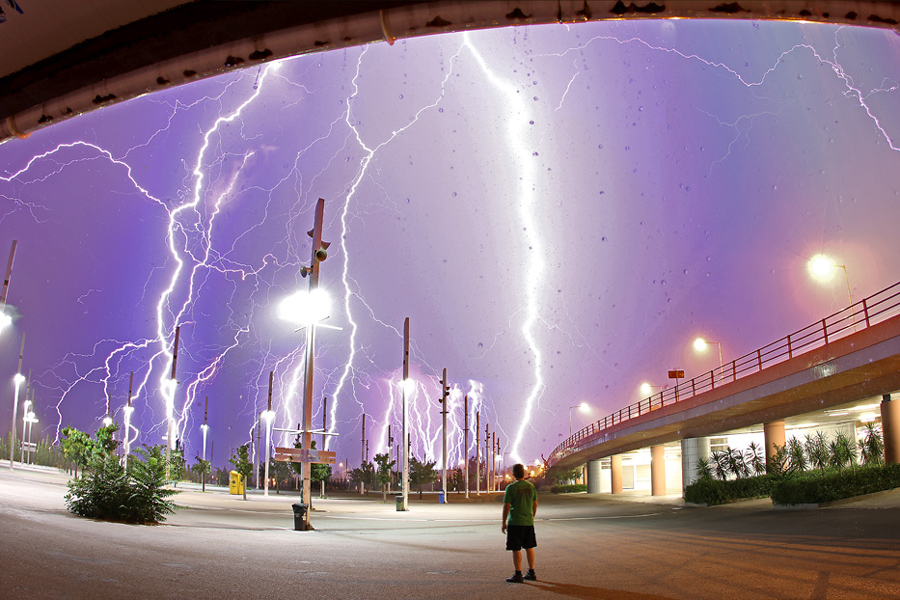 Der Blick geht von unter einer Brücke auf einen Parkplatz mit Lampen und einem violetten Himmel, der von zahllosen gleißenden Blitzen beleuchtet ist. In der Bildmitte steht eine Person, rechts verläuft eine Brücke.