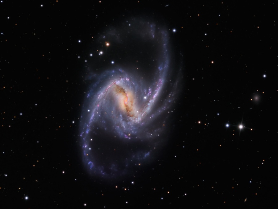 Im Bild leuchtet eine Balkenspiralgalaxie mit dunklen Staubbahnen über den Zentrum und einem aufgefächerten Spiralarm nach rechts oben. Im Hintergrund leuchten wenige Sterne.