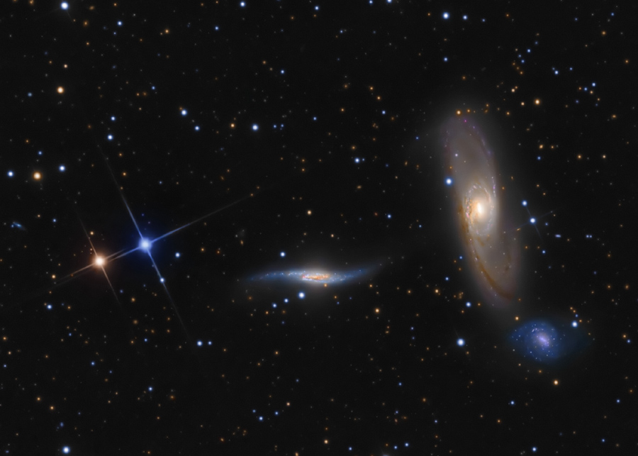 Im Bild sind zwei größere Galaxien, rechts eine "stehende", schräg sichtbare Spiralgalaxie, in der Mitte eine längliche Spiralgalaxie mit herausgezogenen Armen und rechts unten eine kleine, blasse, blaue Spiralgalaie. Rechts sind zwei hellere Sterne, einer blau, einer orangefarben. Im Hintergrund sind Sterne verteilt.