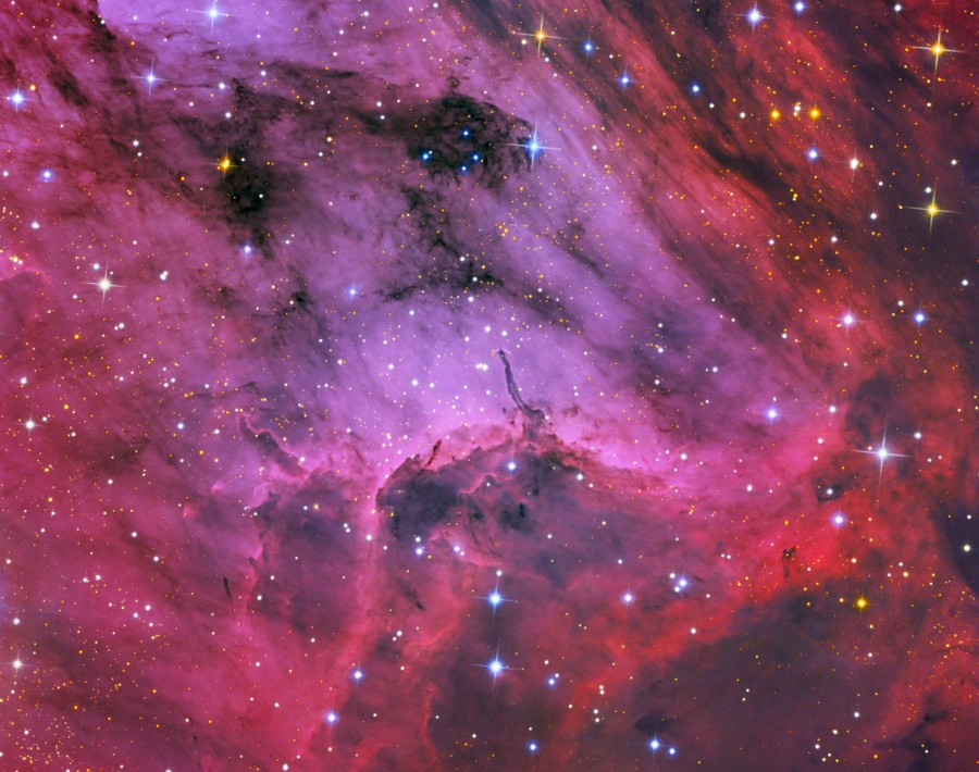 Das Bild zeigt einen magentafarbenen Teppich aus leuchtenden Wolken mit vielen Sternen und einem dunklen Herbig-Haro-Objekt in der Mitte.