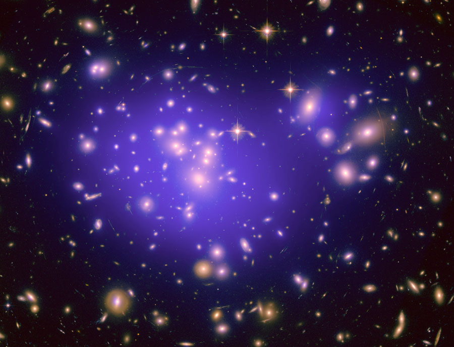 Im Bild sind viele Lichtflecken verteilt, die fast allesamt Galaxien sind. In der Mitte sind sie von einen blauen Nebel umgeben.