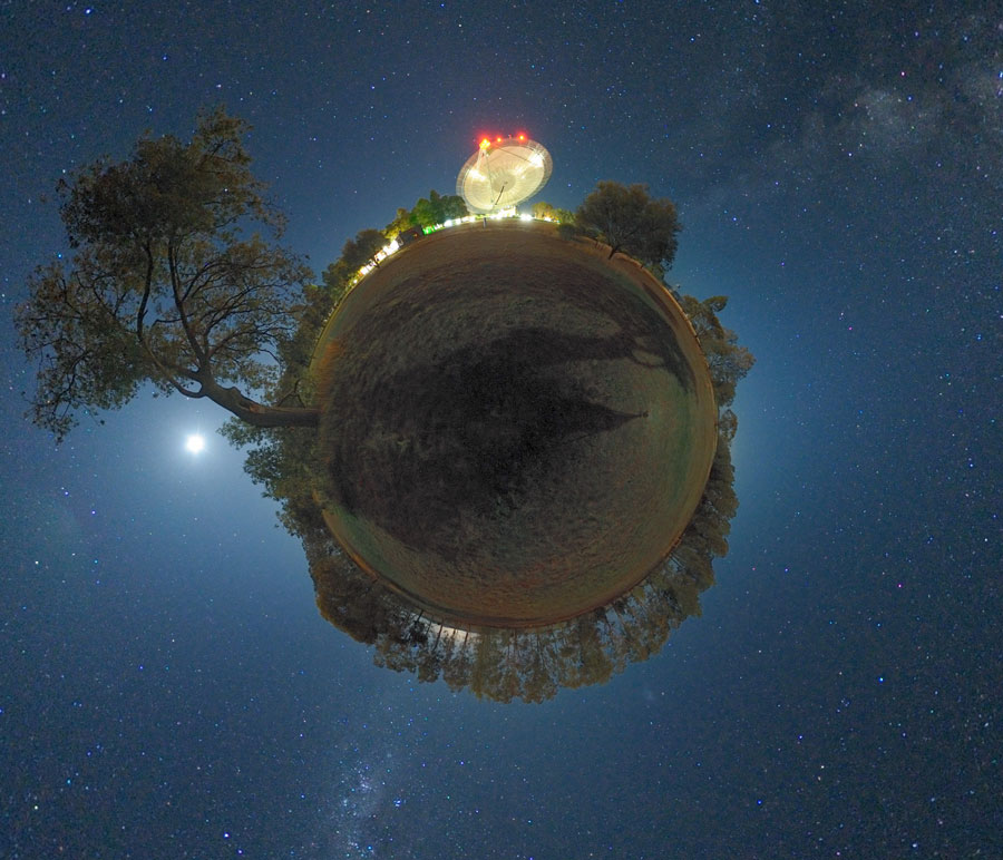 Vor einem blauen Himmel schwebt eine Kugel, links leuchtet unter einem Baum der Mond, am unteren Rand ragt eine Baumreihe am Rand der Kugel auf, und oben in der Mitte steht ein hell beleuchtetes Radioteleskop.