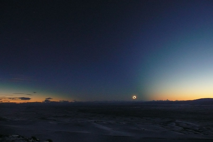 Über der dunklen Erde leuchtet am Horizont die Korona um den Mond, links und rechts ist der Horizont hell, in der Mitte ist der Schatten der Erde am blauen Himmel erkennbar.