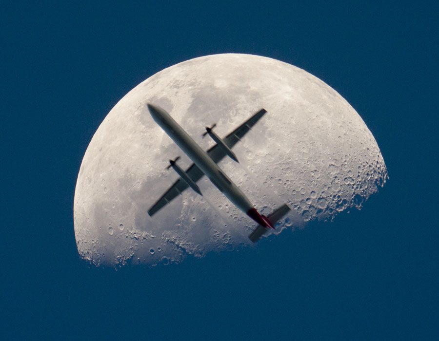 Ein Flugzeug fliegt vor dem Mond vorbei, scheinbar ist es fast halb so groß wie der Himmelskörper. Der Mond ist von oben etwa halb beleuchtet, an der Schattengrenze sind markante Krater erkennbar.