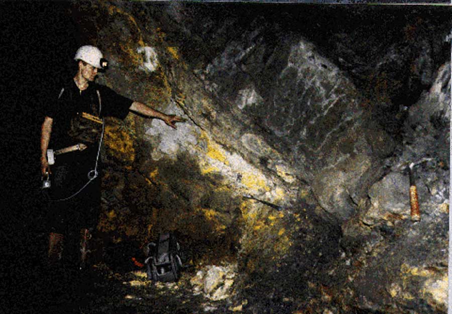 Eine Person mit weißem Helm und schwarzer Kleidung zeigt in einem Stollen auf Spuren, die ein natürlicher Kernreaktor hinterlassen hat.