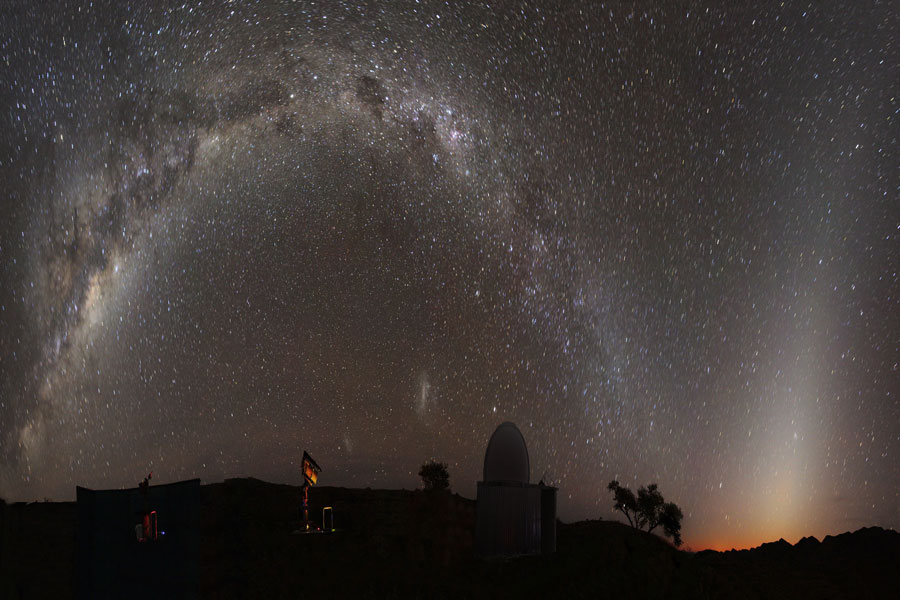 Das Bild ist horizontal verkürzt. Links wölbt sich die Milchstraße fast über das ganze Bild, rechts daneben steigt ein Lichtdreieck auf, es ist das Zodiakallicht. Am Horizont ist die Silhouette einer Sternwarte und ein rot beleuchtetes Fernrohr erkennbar.