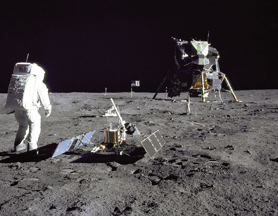 Der Apollo-Astronaut Edwin "Buzz" Aldrin steht auf dem Mond neben dem soeben aufgebauten Mondseismometer und blickt zur Landefähre Eagle. Der Himmel ist schwarz, der Boden von grauem Staub bedeckt, in dem sich Fußspuren der Astronauten abzeichnen.