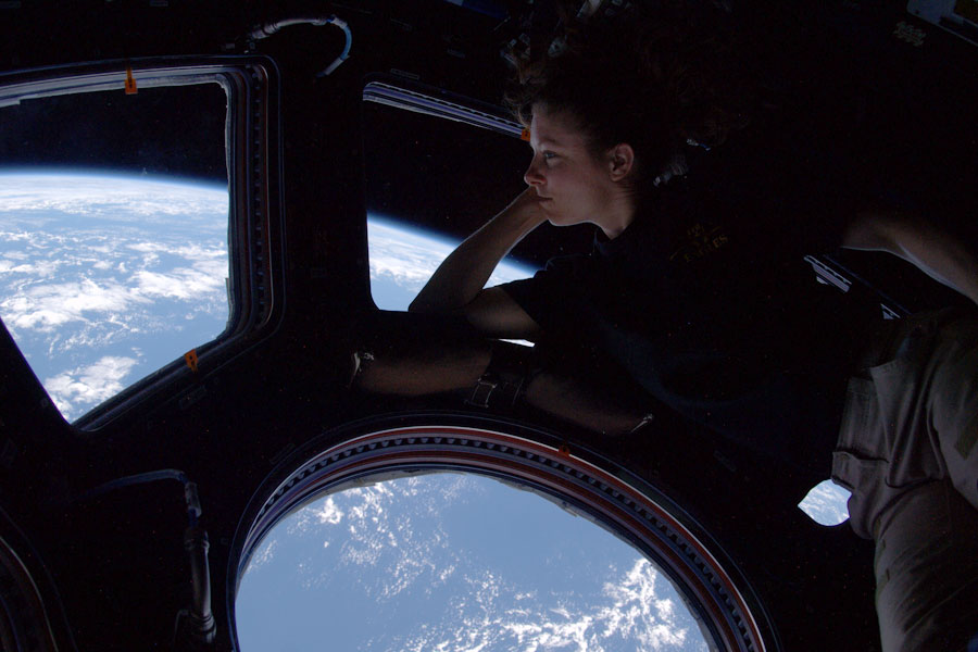 Unten ist durch das Fenster der Cupola auf der ISS die Erde mit blauen Meeren und weißen Wolken zu sehen, eine Astronautin blickt nachdenklich hinaus.