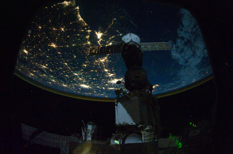 Hinter der Raumstation mit einer angedockten Sojus-Kapsel und Solarpaneelen leuchten auf der Erde die Nachtlichter der USA.