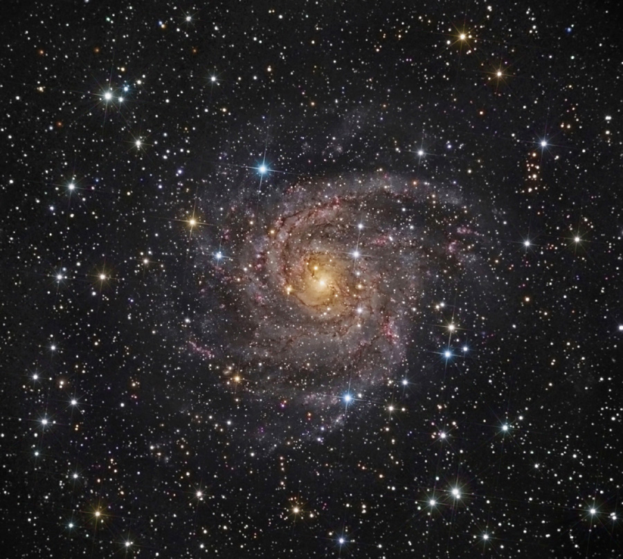 Hinter dicht gestreuten, verschieden großen Sternen in unterschiedlichen Farben schwebt eine von oben sichtbare Spiralgalaxie mit ausgeprägten, losen Spiralarmen und einem gelb leuchtenden Kern.