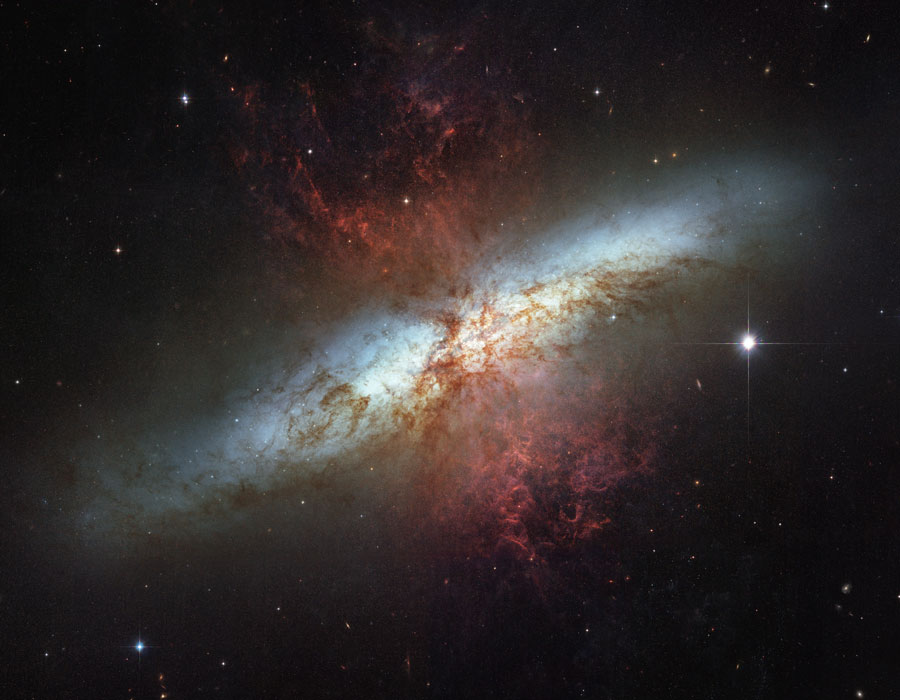 Die verworrene, weißliche Galaxie veräuft von links unten diagonal durchs Bild, in der Mitte ist sie von rot leuchtenden Wolken überzogen.