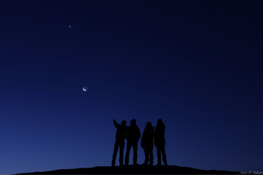 Vier dunkle Silhouetten stehen vor einem sehr dunklen blauen Himmel, die rechte Person hebt die Hand zum Sichelmond, darüber leuchtet der Planet Venus.