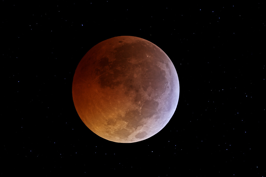 Mitten im Bild leuchtet ein rötlich gefärbter Mond, der nach links unten hell und leicht bläulich wird. Die dunklen Flecken der Lavameere sind gut erkennbar.