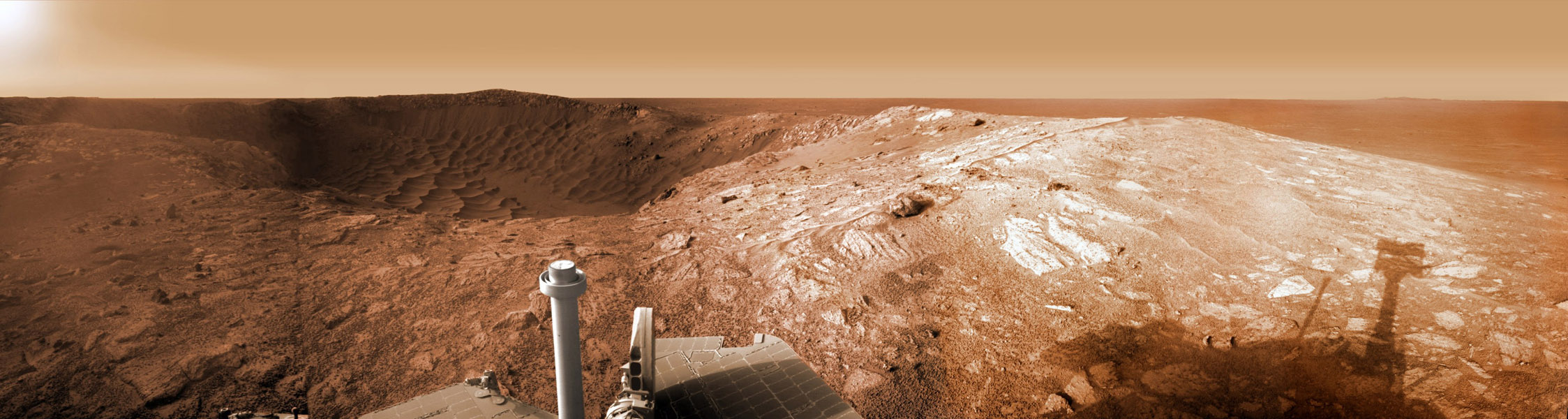 Panorama der Marsoberfläche, in der Mitte ragen unten Teile des Rovers auf, links am Horizont ist eine dunkle Stelle, rechts ein Hügel mit hellen Steinen.