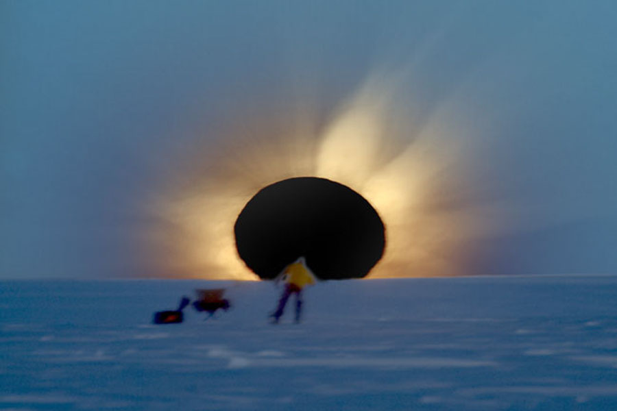 Hinter einer verschneiten Ebene geht die Sonne unter, sie ist vom Mond verdeckt, der einen schwarzen Kreis bildet. Rund um den Mond leuchten die Strahlen der Korona. Vor dem Mond befindet sich eine Person in einiger Entfernung.