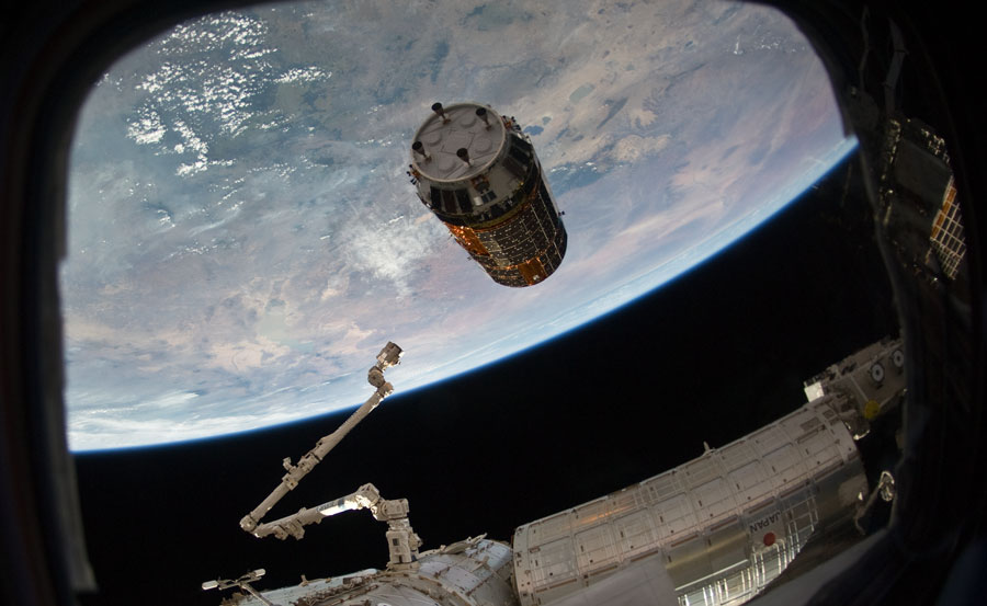Durch das Fenster der Internationalen Raumstation fällt der Blick auf die Erde, davor schwebt eine zylinderförmige Raumsonde. Ein Greifarm wird in ihre Richtung geschwenkt, um sie einzufangen.