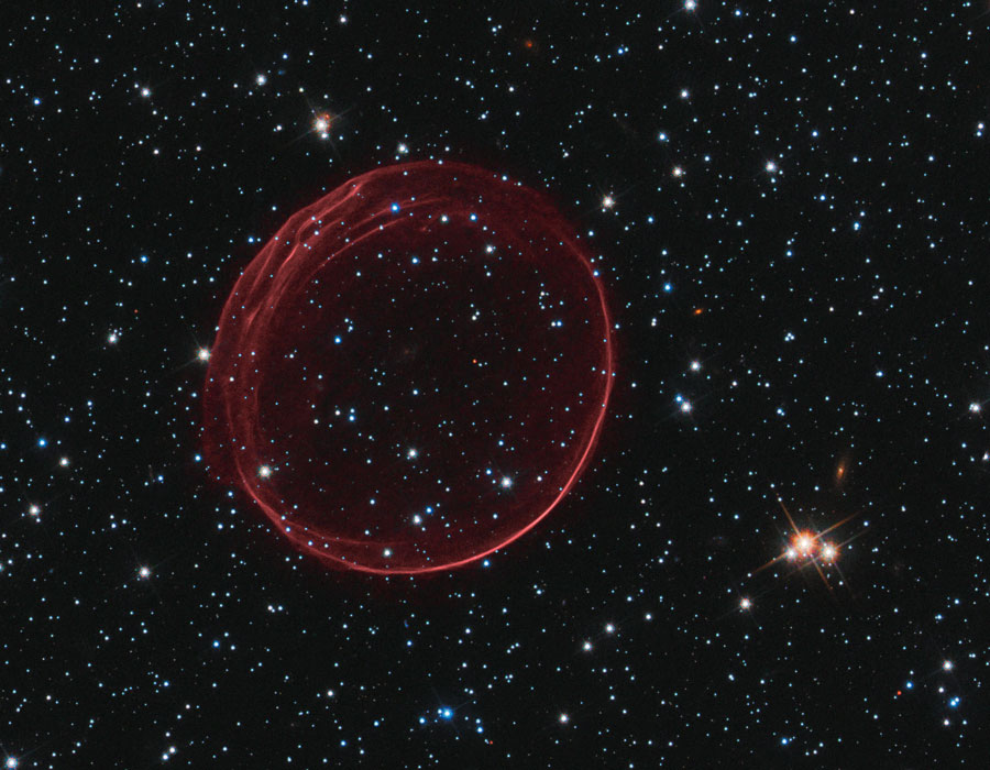Vor einem Hintergrund aus zarten Sternen zeichnet sich eine ebenso zarte rote Blase ab, man sieht nur ihren Umriss.