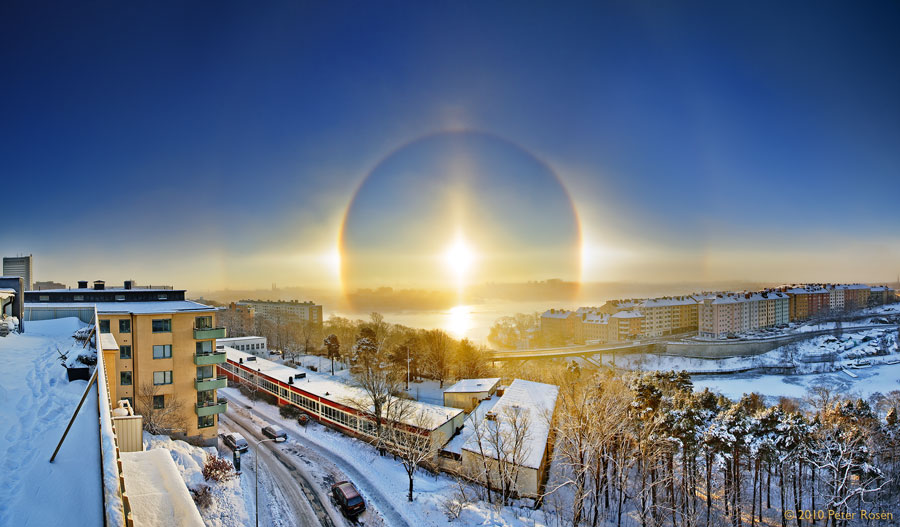 Am Horizont hinter einer verschneiten Kleinstadt leuchtet eine Sonne mit eindrucksvollen Ringen, Lichtsäulen und Nebensonnen am blauen Morgenhimmel.