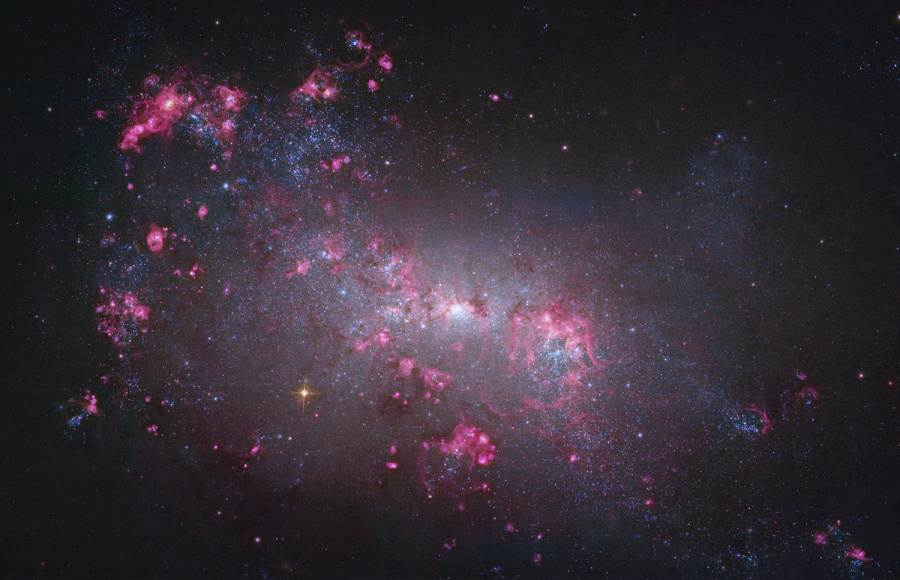 Vor einer weiß leuchtenden Sternansammlung sind magentafarben leuchtende Nebel verteilt, sie wirken wie Wolkenfetzen. Die Nebel sind links und in der Mitte, die rechte obere Bildecke ist dunkel.