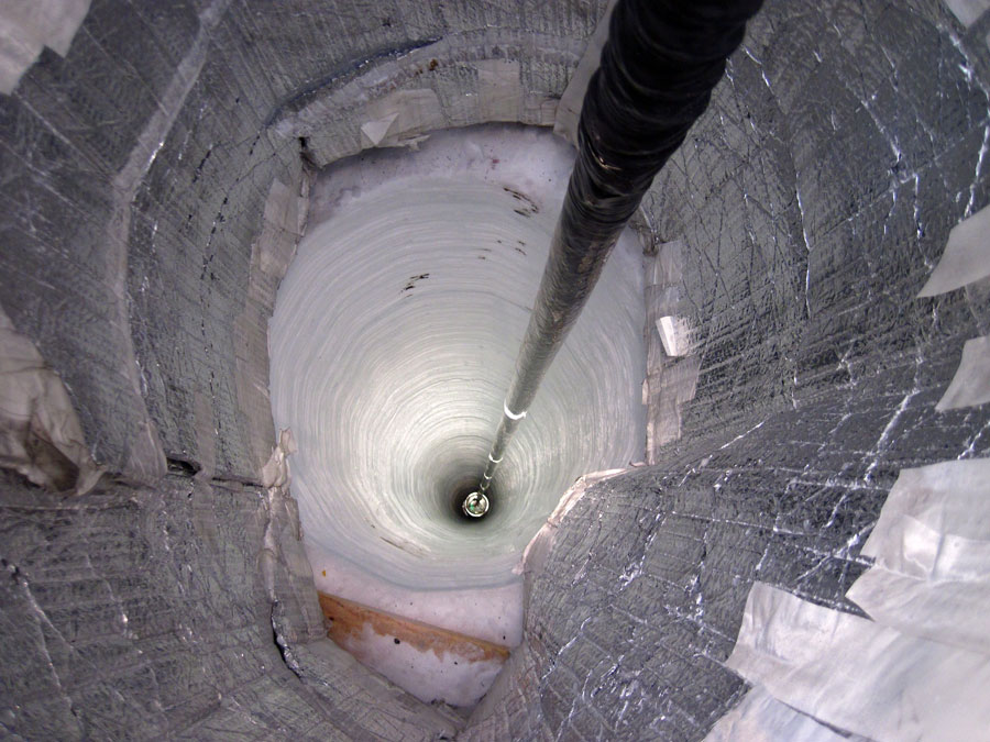 Das Bild zeigt den Blick in ein Bohrloch, in dessen Mitte eine lange Stange verschwindet. Das Schmelzloch führt tief ins Eis und ist oben mit silbrig reflektierendem Material verkleidet.