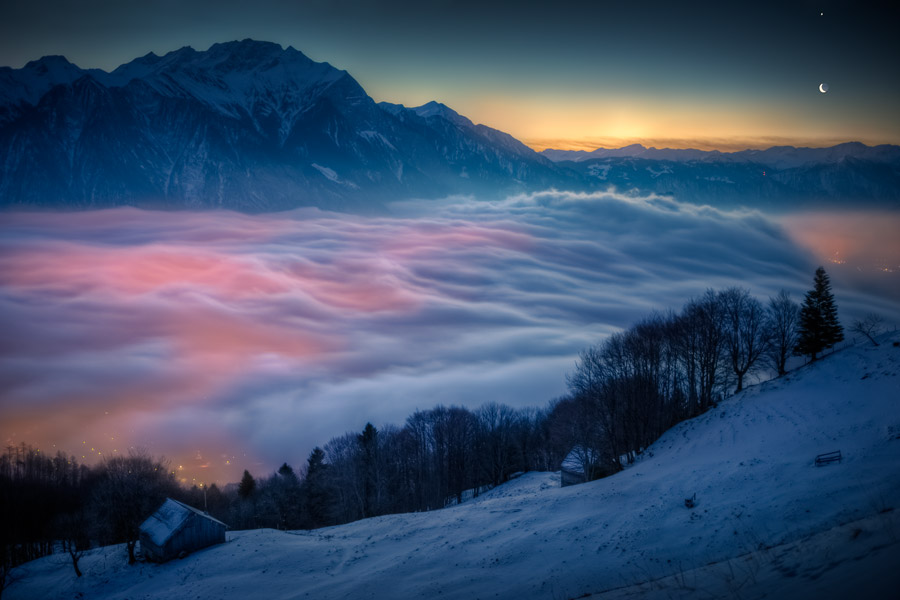 Der Blick fällt über einen verschneiten Hang hinab auf eine Nebeldecke, die von unten beleuchtet wird. Dahinter ragt links ein Gebirge auf. Rechts leuchtet ein Sichelmond in der Morgendämmerung.