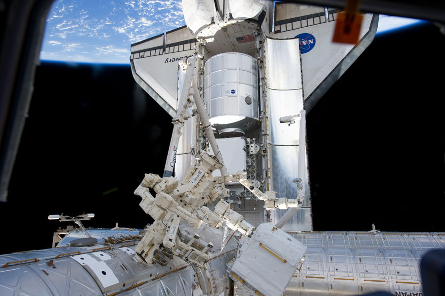 Die Raumfähre ist mit der Nase nach unten an die Raumstation gekoppelt. Vor der geöffneten Ladebucht warten der Canadarm2 und Dextre. Dahinter schwebt oben die Erde im Weltraum.