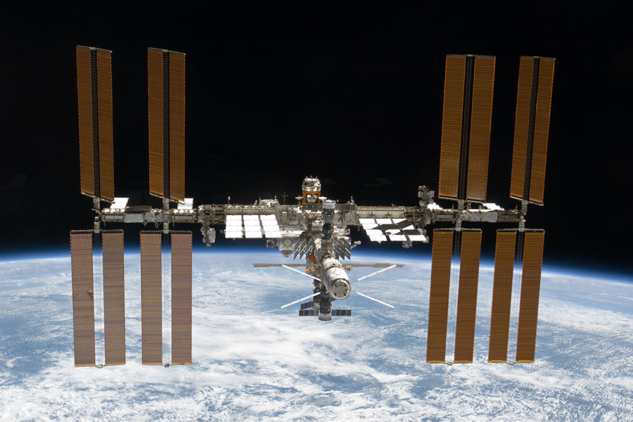 Über der Erde schwebt die Internationale Raumstation. Links und rechts sind riesige Solarpaneele angebracht.